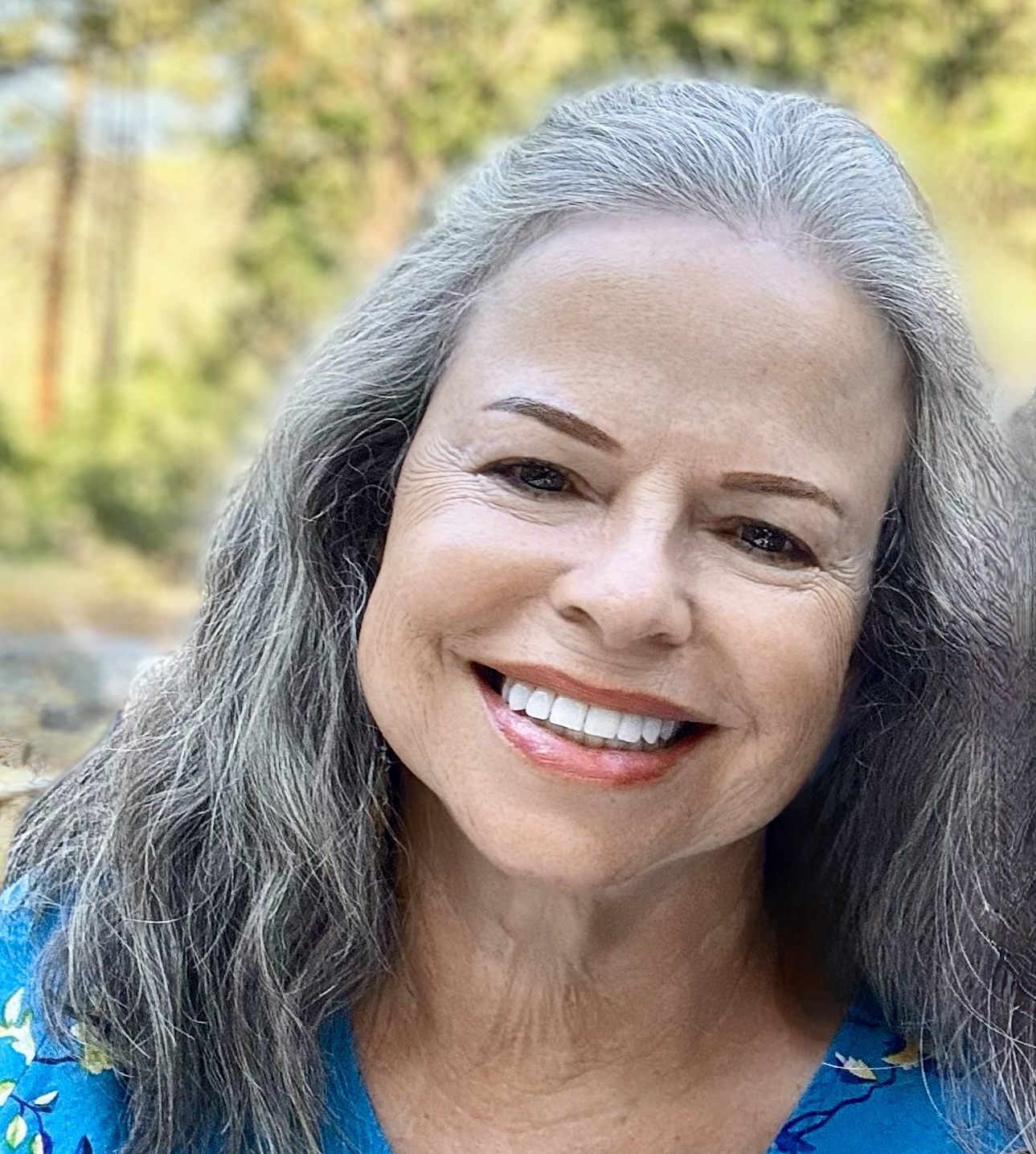 Headshot of Gale O’Brien smiling outdoors, with long gray hair and wearing a bright blue floral top against a softly blurred natural background.