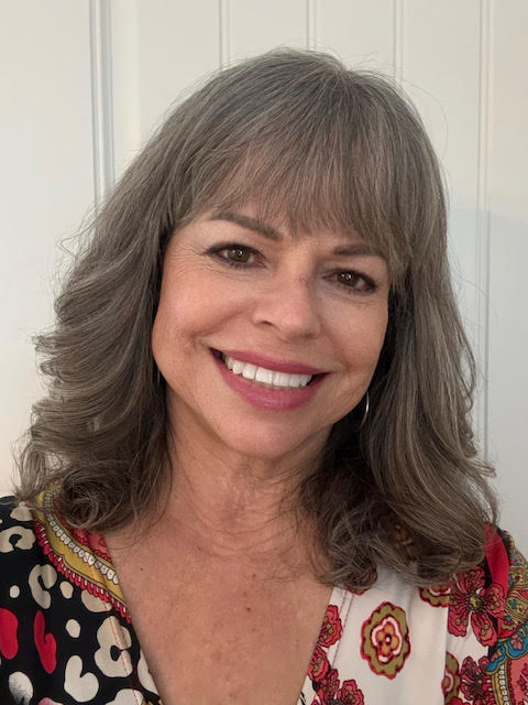 Headshot of a smiling woman with shoulder-length gray hair and bangs, wearing a patterned V-neck top, against a light paneled background.