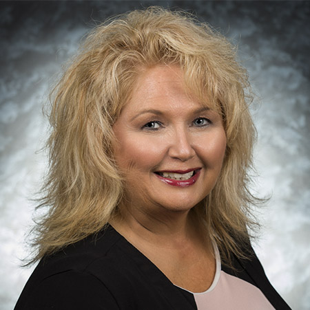 Portrait of Pamela Duran, a smiling blonde woman wearing a black and blush-pink top against a soft blue-gray background.